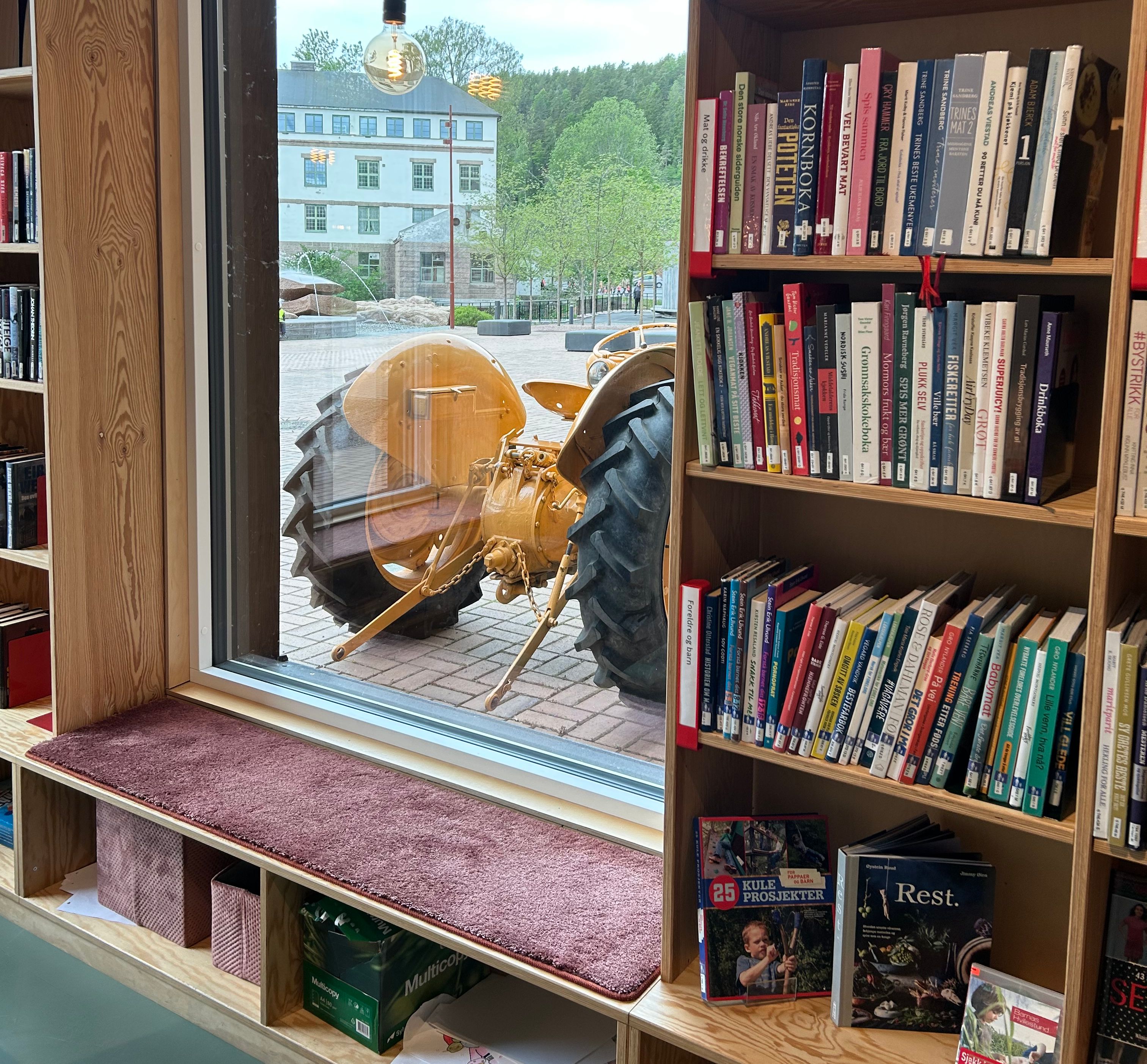 et bibliotek med plassbygde reoler og et vindu med en traktor utenfor