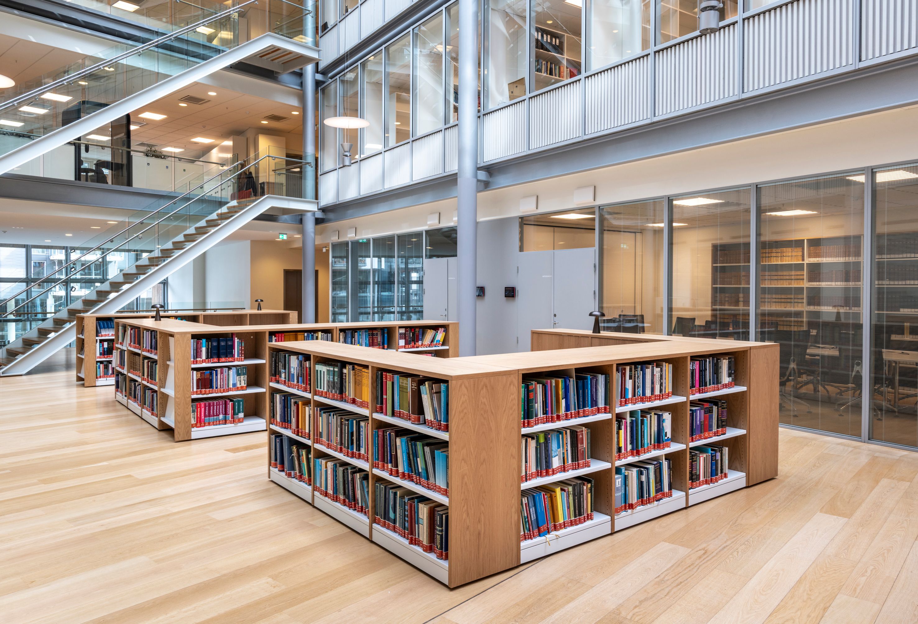 A modern library interior with light wooden floors, low wooden bookshelves, and a contemporary glass-railed staircase.