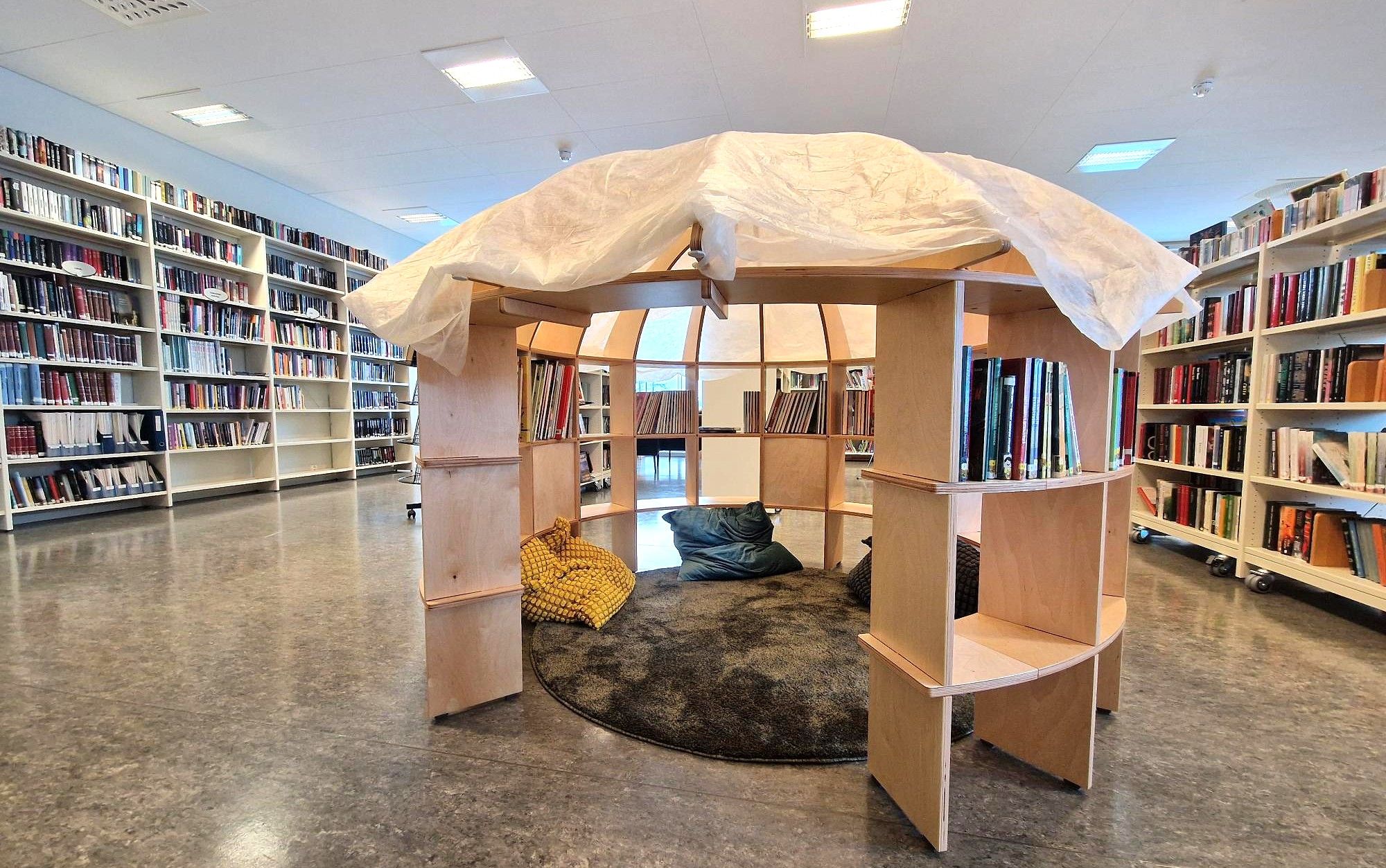 A library with a central wooden reading nook featuring a fabric roof, soft seating, and surrounding tall bookshelves.