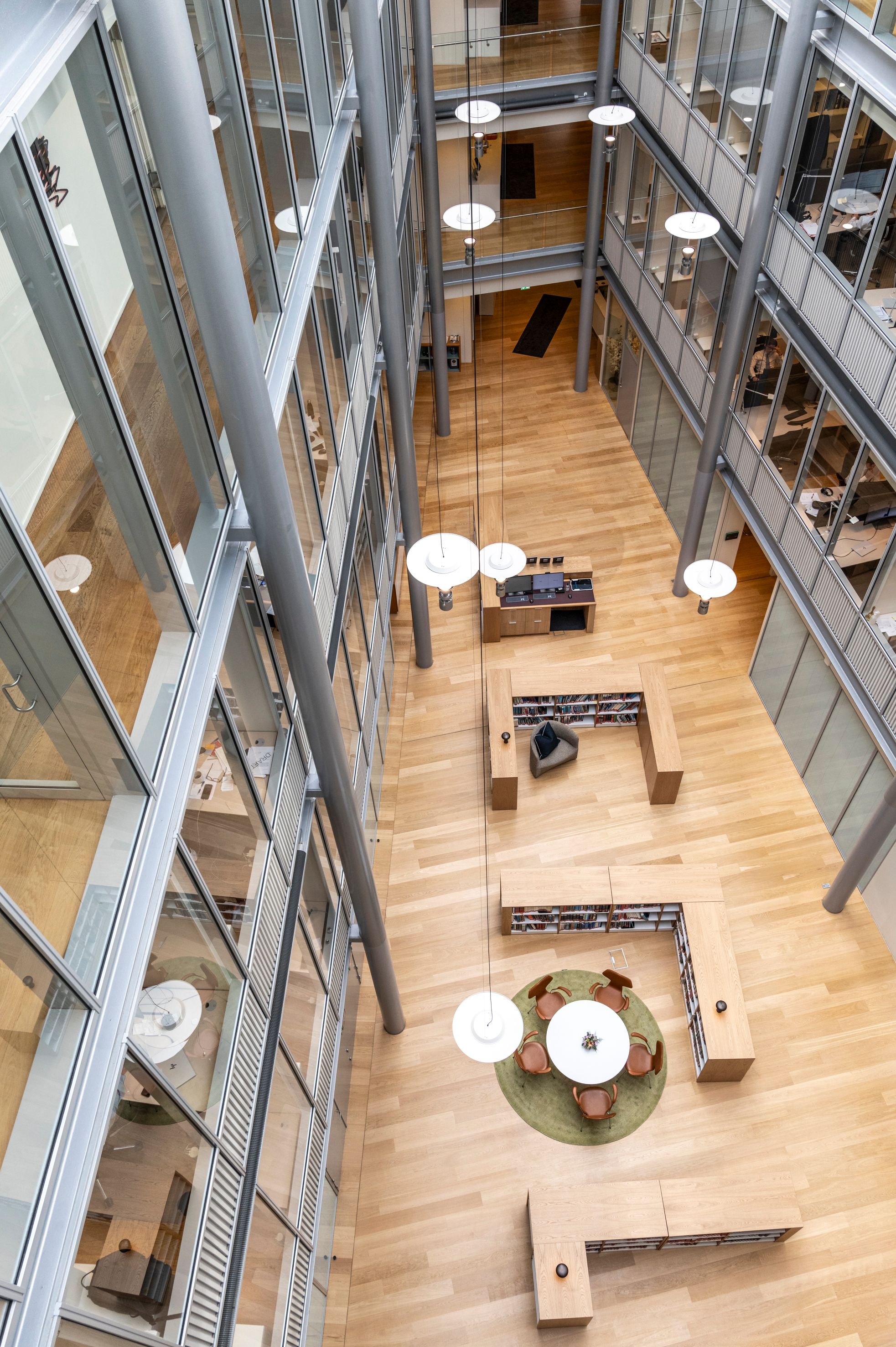 Overhead view of a modern multi-story building atrium with glass-walled offices, light wood floor, modern furniture, and hanging pendant lights.