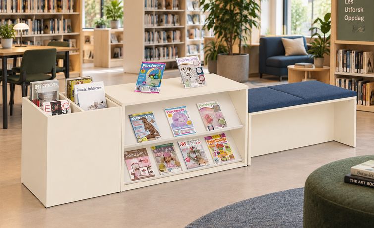 A modern library lounge area with a beige modular magazine display unit, an attached bench with blue cushions, and bookshelves in the background.