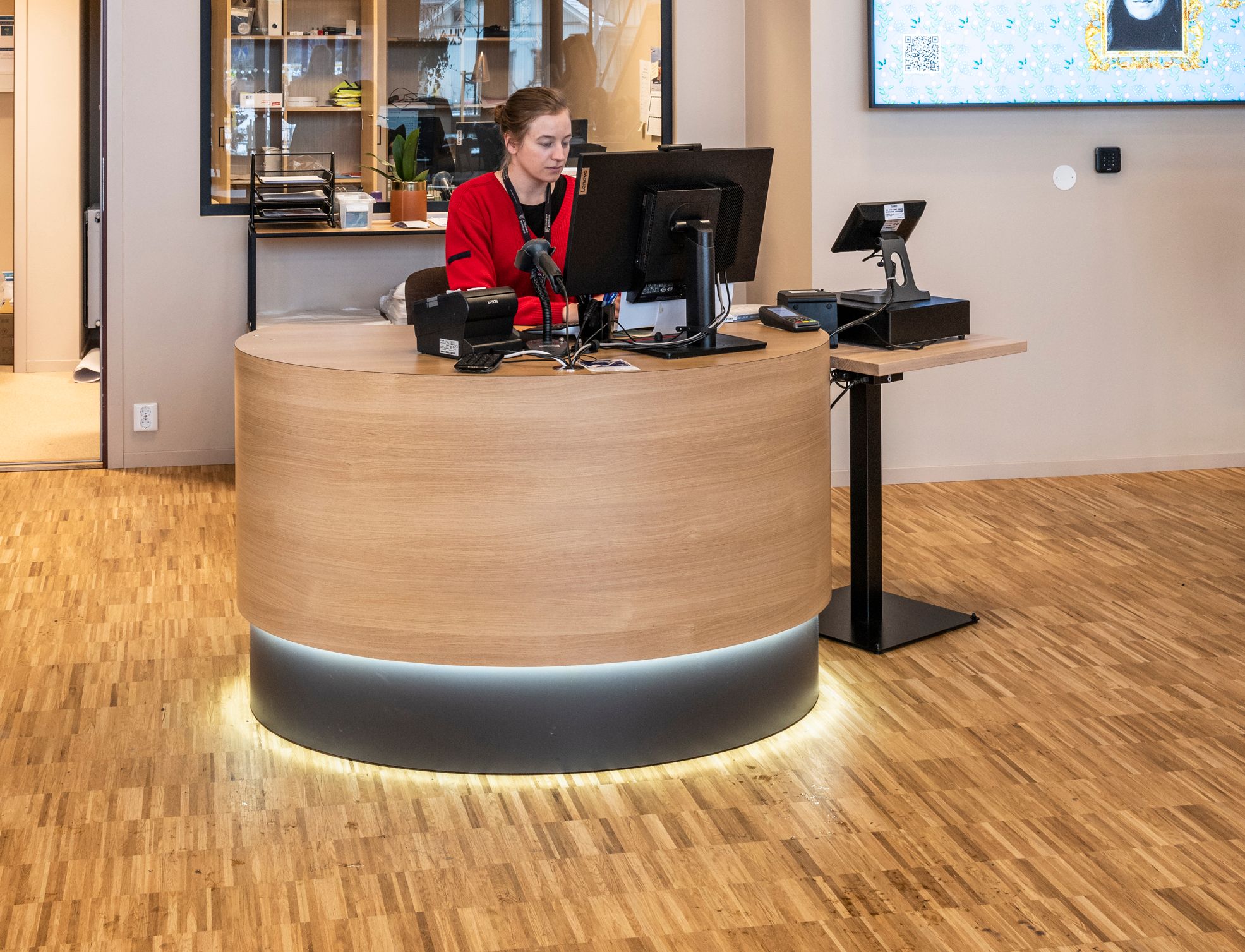 en kvinne i en rød jakke sitter ved et skrivebord med en datamaskin