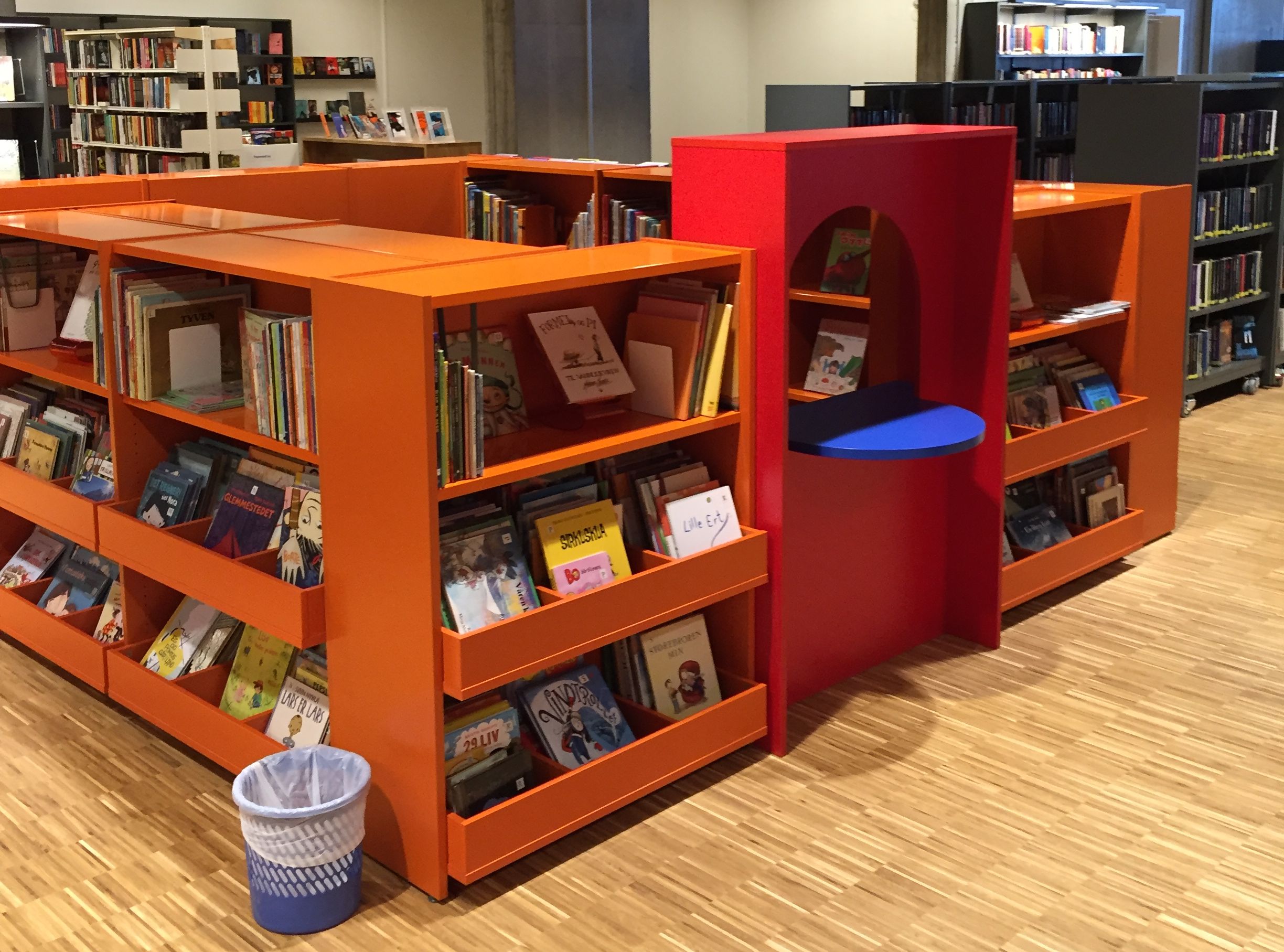 et bibliotek med oransje hyller fylt med bøker og en søppelbøtte.