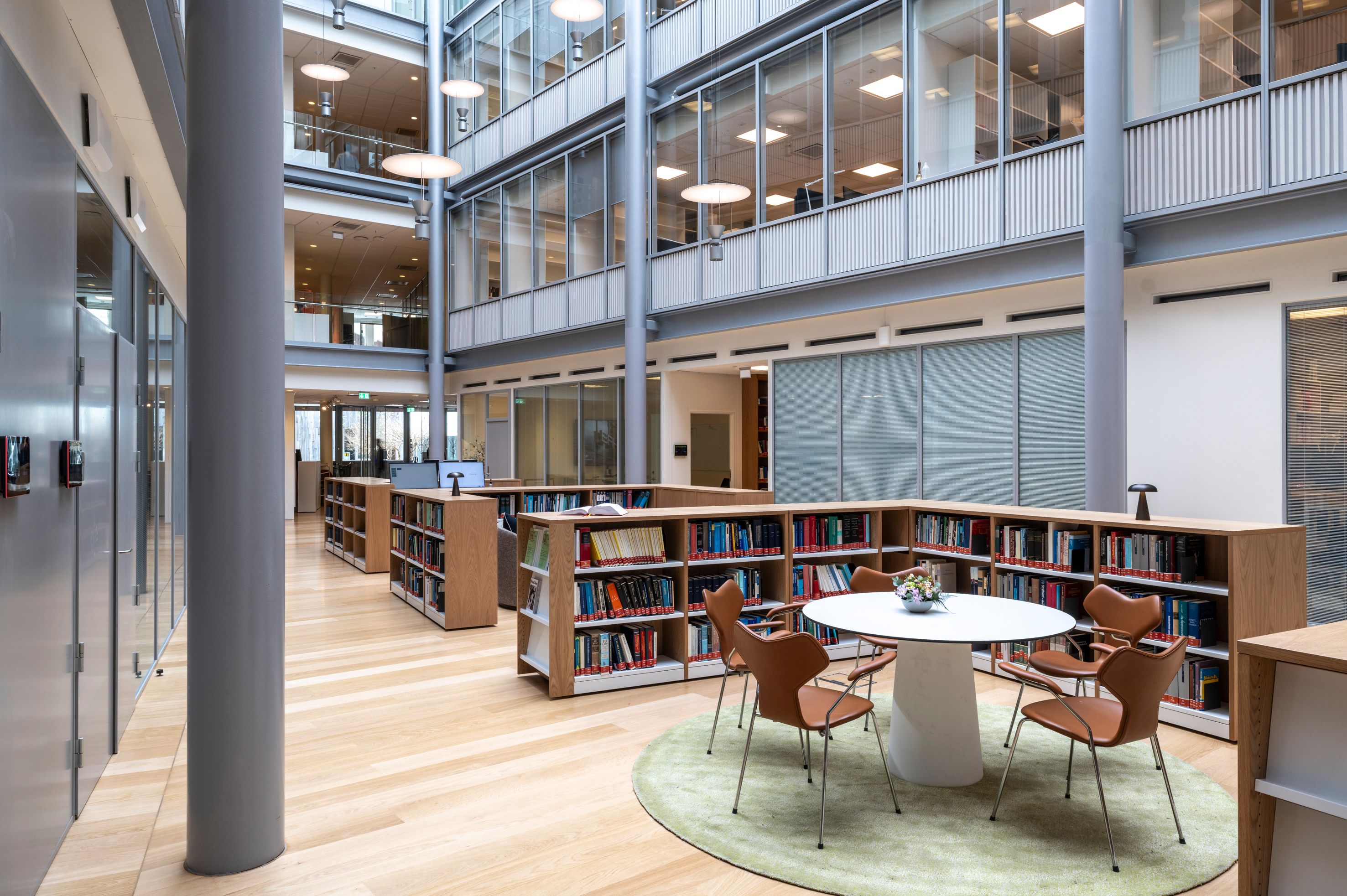 Modern, multi-story library with an open atrium, bookshelves, and a central white table with four brown chairs on a green rug.