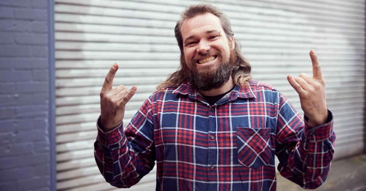 A man with long hair and a beard is wearing a plaid shirt and making a horns sign .