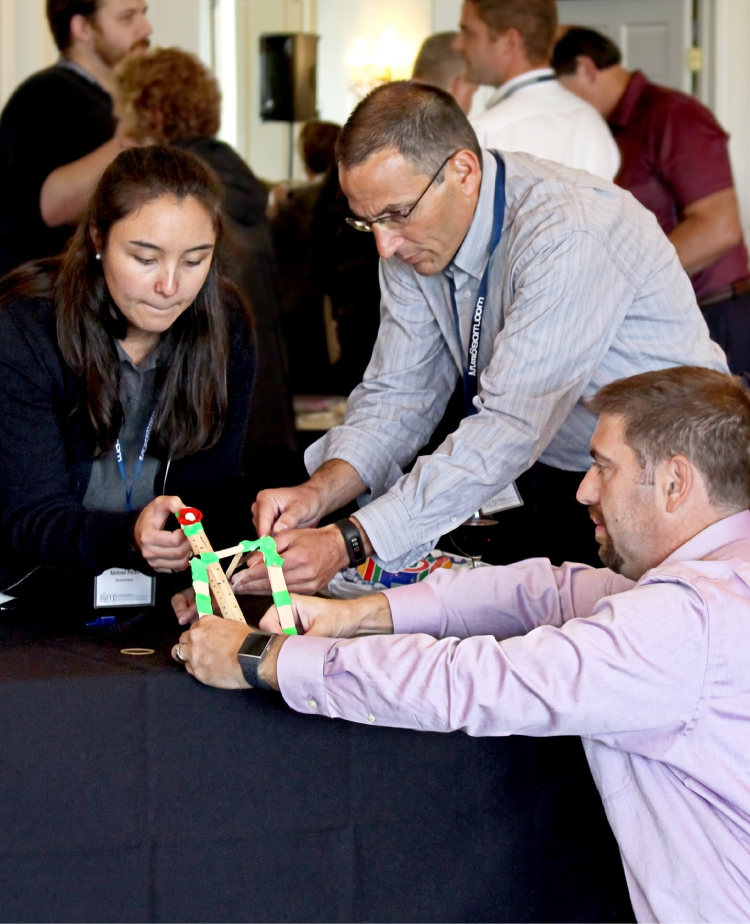 Three participants building a catapult from craft sticks and rubber bands at a workshop table