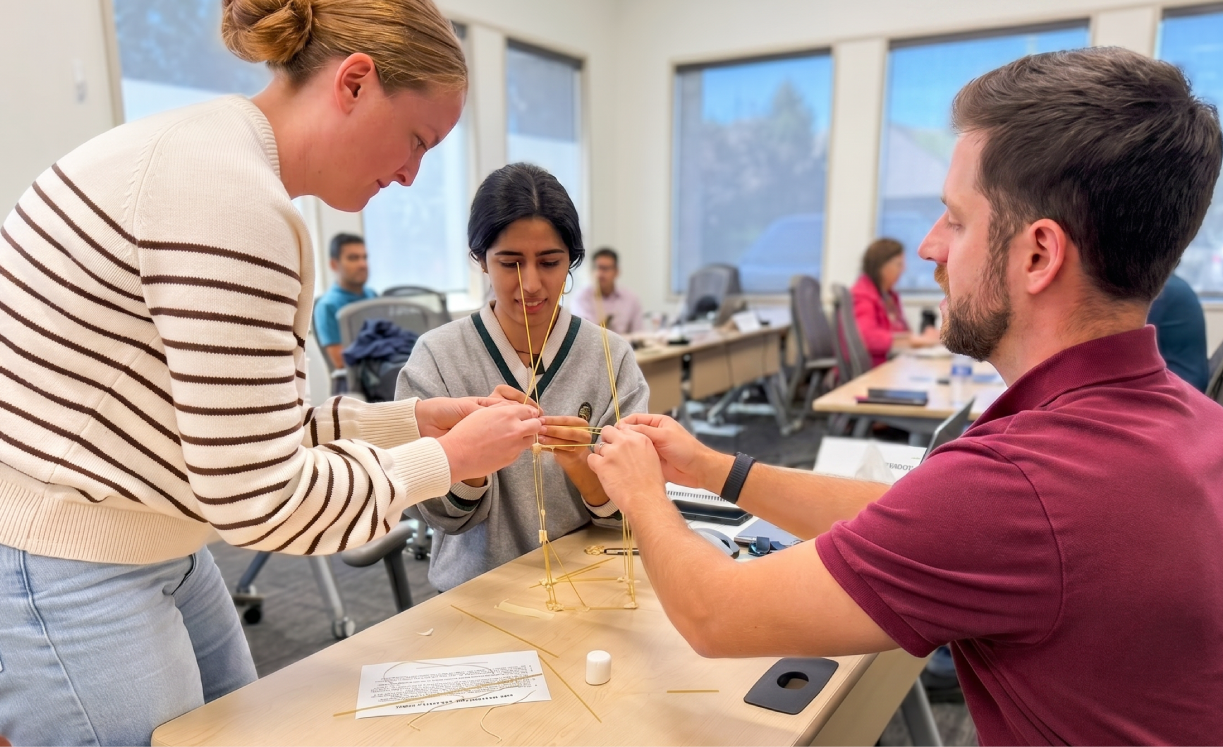 Three workshop participants inspecting a small spaghetti-and-tape structure at a classroom table