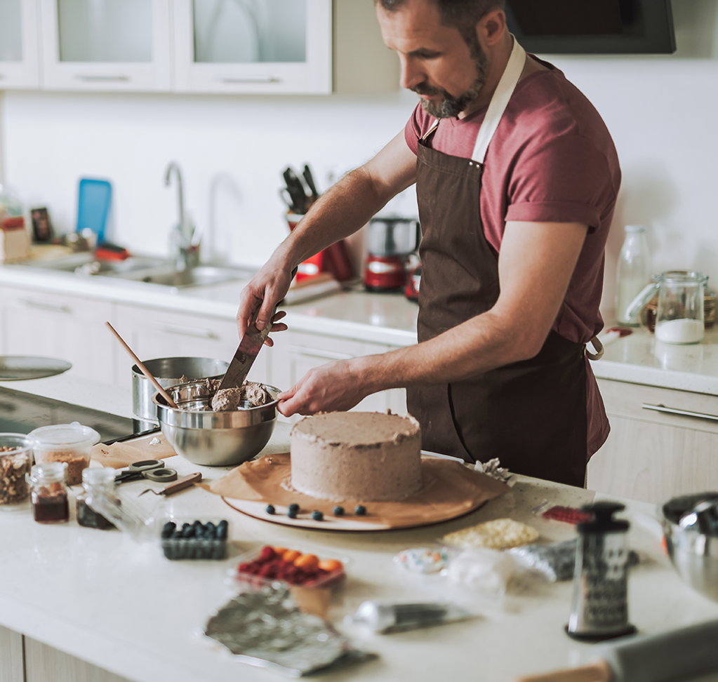 Man wearing an apron frosting a cake with ingredients surrounding him.