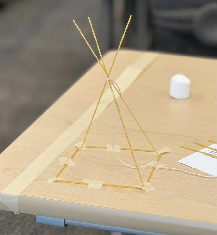A completed spaghetti tower taped to a table beside a marshmallow and leftover pasta