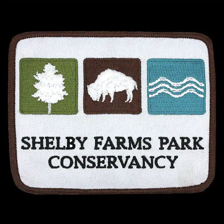 Rectangular embroidered patch for Shelby Farms Park Conservancy with a tree, a bison and water in three coloured squares above black lettering, finished with a brown merrowed border.