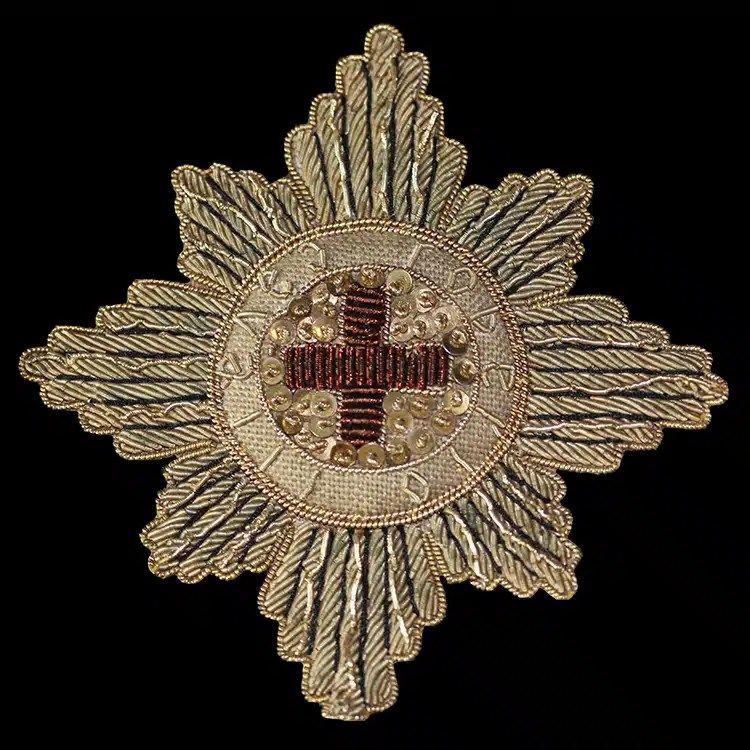 Eight-pointed star badge worked in antique gold bullion wire, with sequins and a red wire cross at the centre, on black.