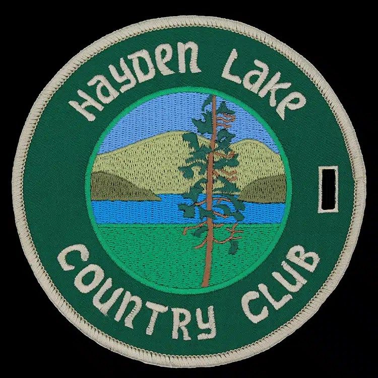 Round embroidered bag tag with a lake, hills and a tall pine in the centre, a green ring with cream lettering and a strap slot.