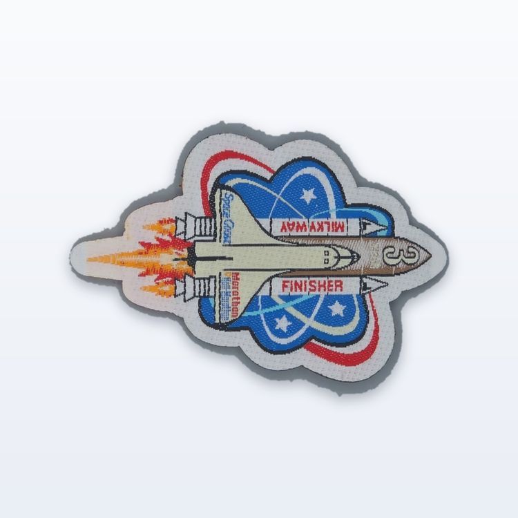 Woven Space Coast Marathon finisher patch shaped like a space shuttle over a blue orbit emblem, cut to its outline.