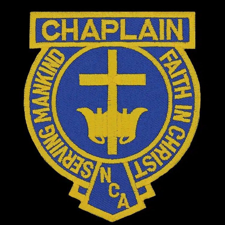 Embroidered shoulder patch in royal blue and gold: a gold cross over a lamp on a blue circle, a title banner across the top and a motto around the ring.