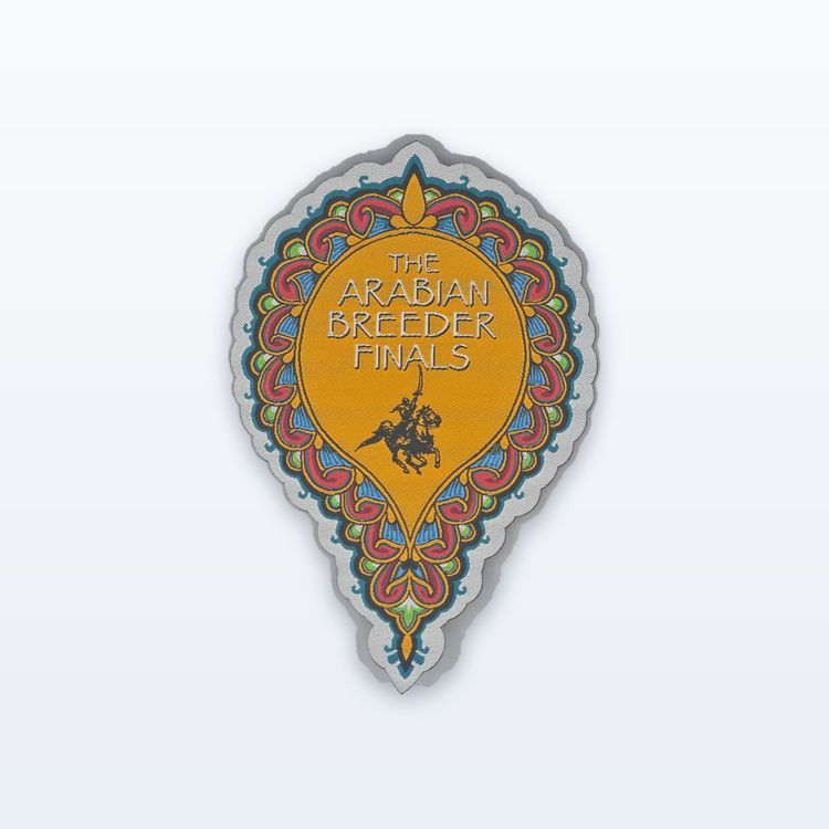 Woven teardrop patch for The Arabian Breeder Finals with an ornate red, blue and green scroll border and a small horse and rider, cut close to the outline.