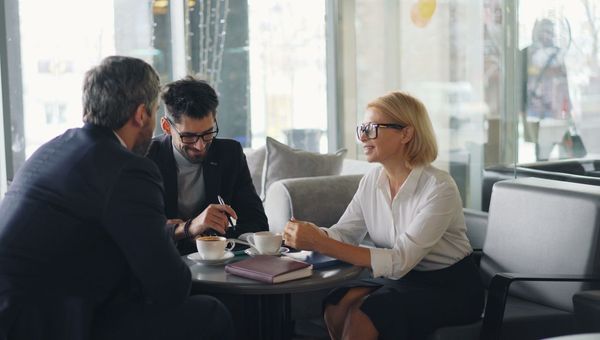Berater im Gespräch mit zwei Kunden an einem Tisch, vor ihnen ein Notizbuch und Kaffee
