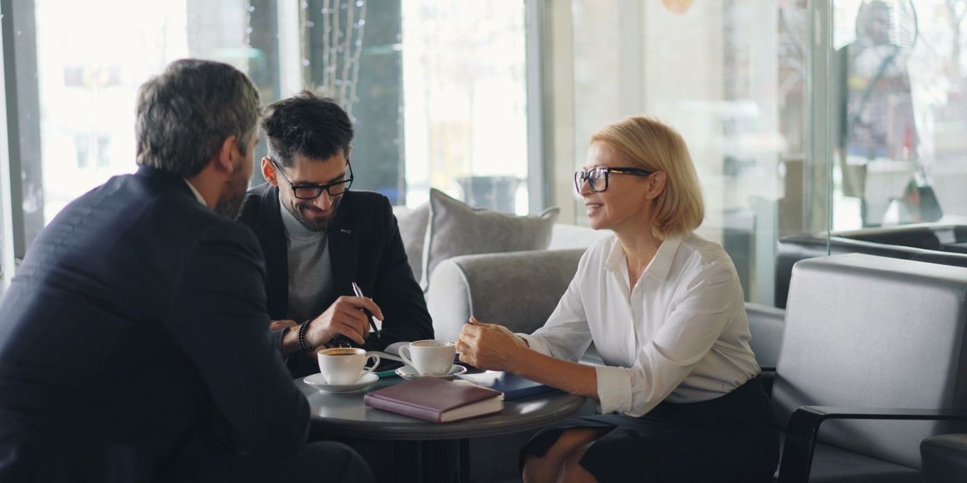 Consultant en rendez-vous avec deux clients autour d'une table, carnet et cafés posés devant eux