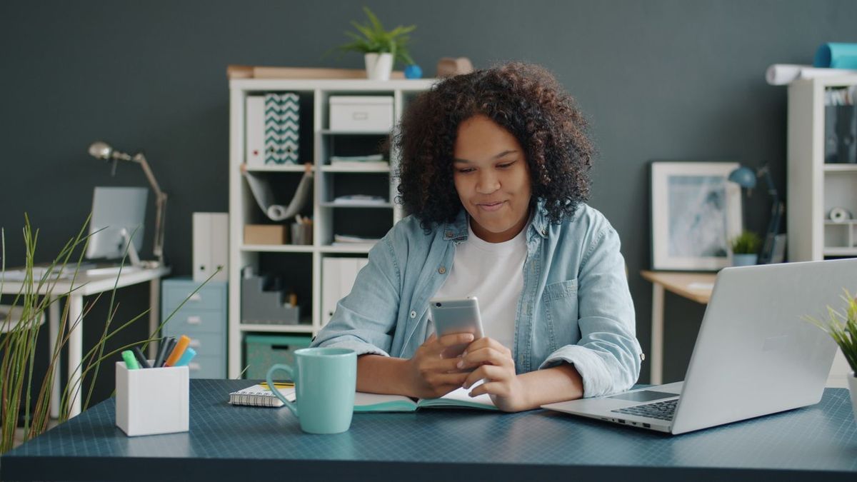 Frau prüft an ihrem Schreibtisch eine Antwort auf dem Smartphone, daneben ein aufgeklappter Laptop