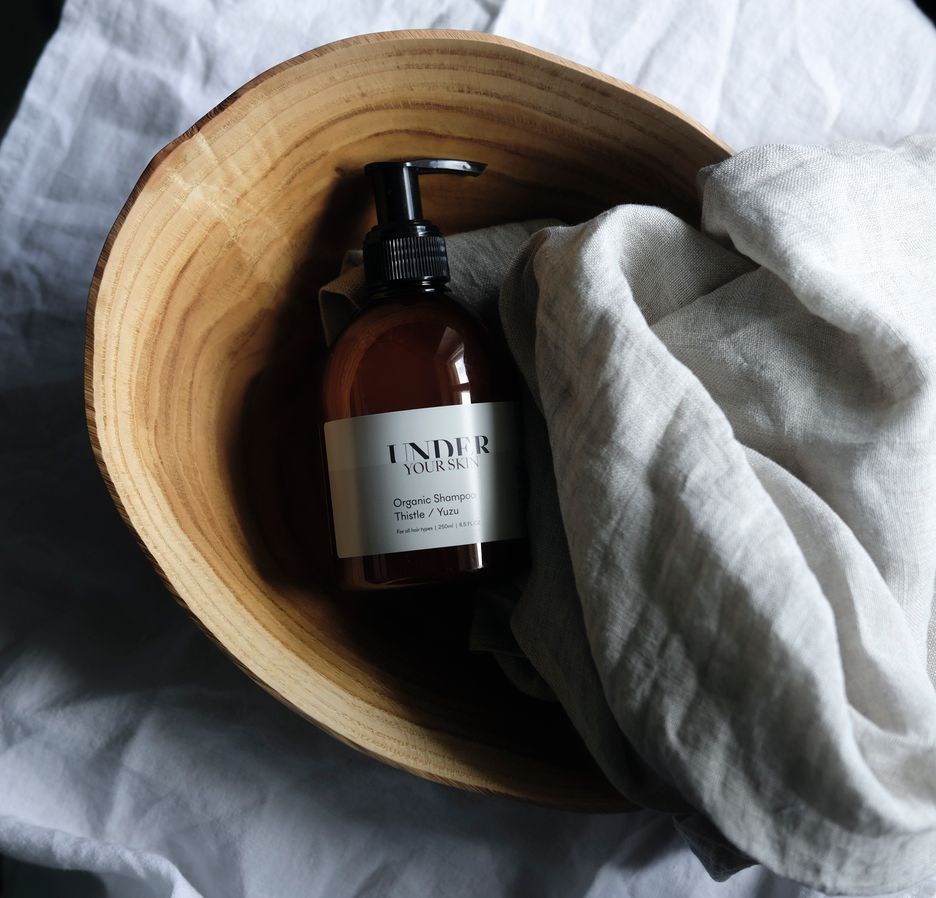 Under Your Skin Organic Shampoo bottle sitting in a wooden bowl with a linen blanket