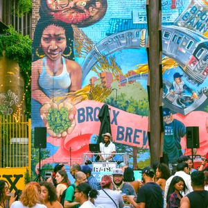 People sitting outside at picnic tables next to a mural of a woman standing next to a subway train and a banner that says Bronx Brewery