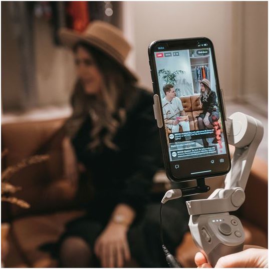 Woman with a black outfit and brown hat sitting on a couch in the background, with a phone on a tripod capturing video footage in the foreground