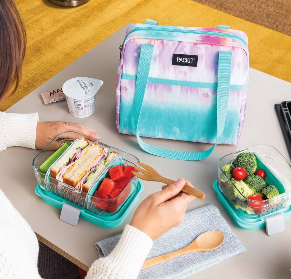 PackIt freezeable lunch bag sitting on a desk with a bento container of a sandwich and fruit and a second bento container of broccoli and tomatoes. Woman is eating lunch with a bamboo fork.
