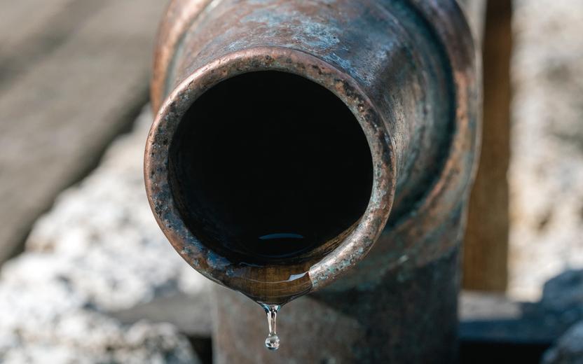 A single, tarnished copper pipe with a slow drip of water forming a bead at its lowest point