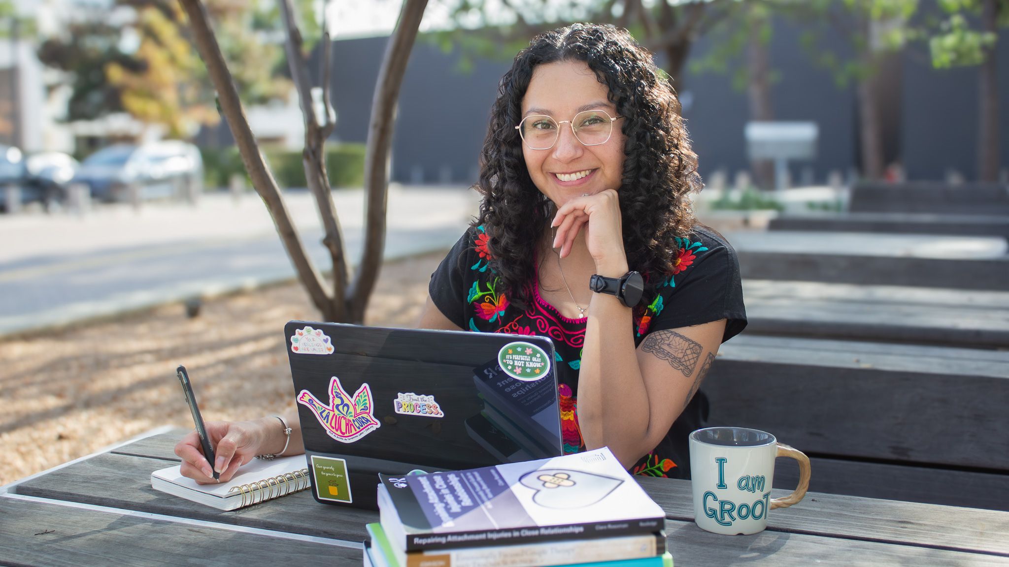 Priscilla Dominguez smiling at an outdoor table with her laptop, books, and a notebook