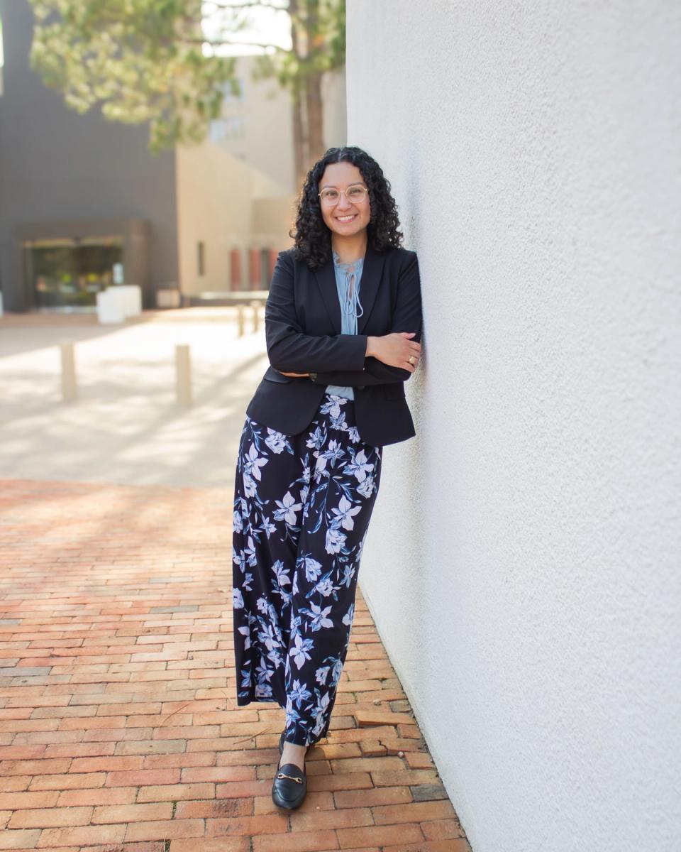 Priscilla Dominguez in a black blazer, leaning against a white wall and smiling