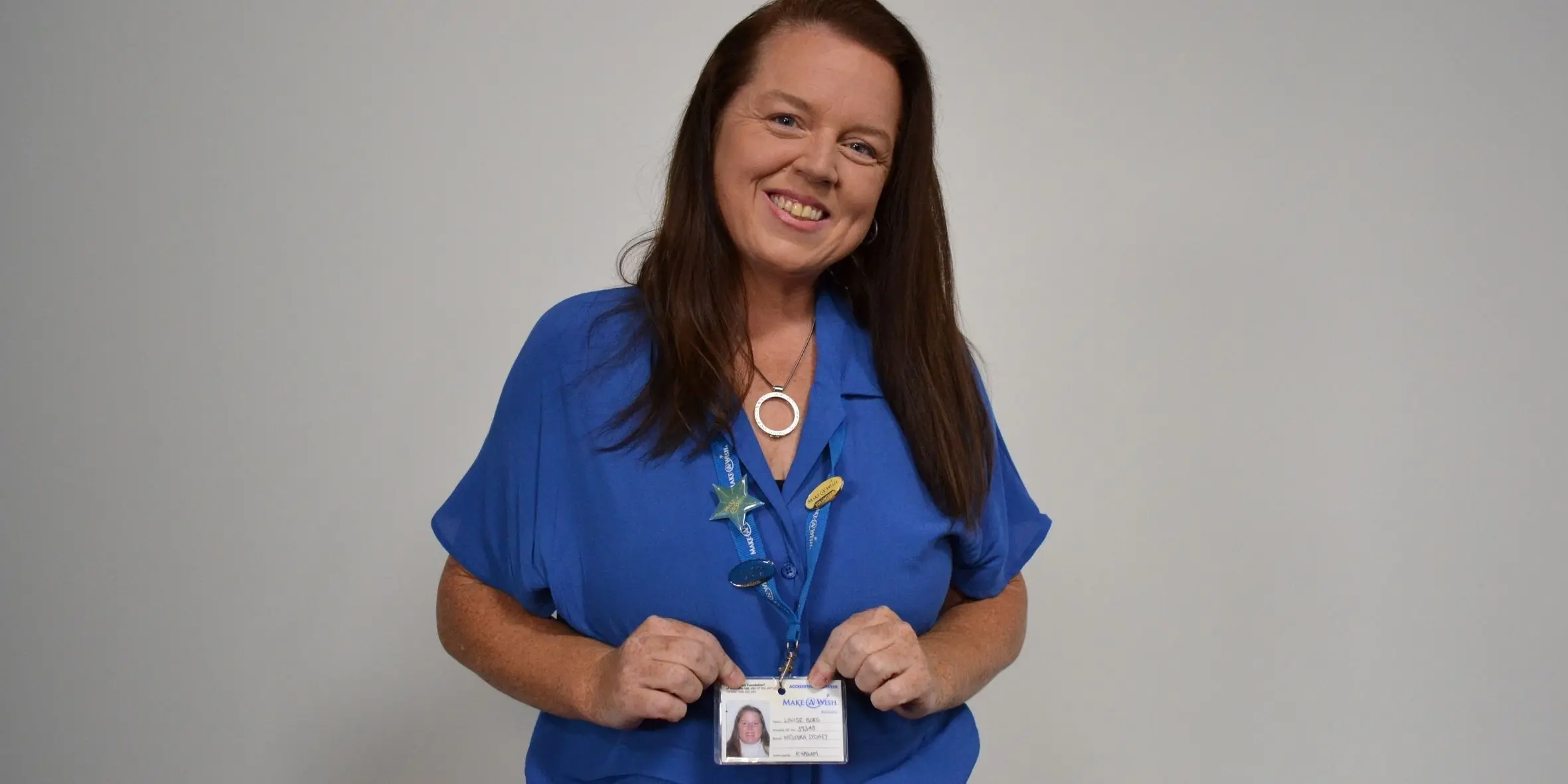 Louise smiling to camera in a blue top and holding her make-a-wish volunteer ID card