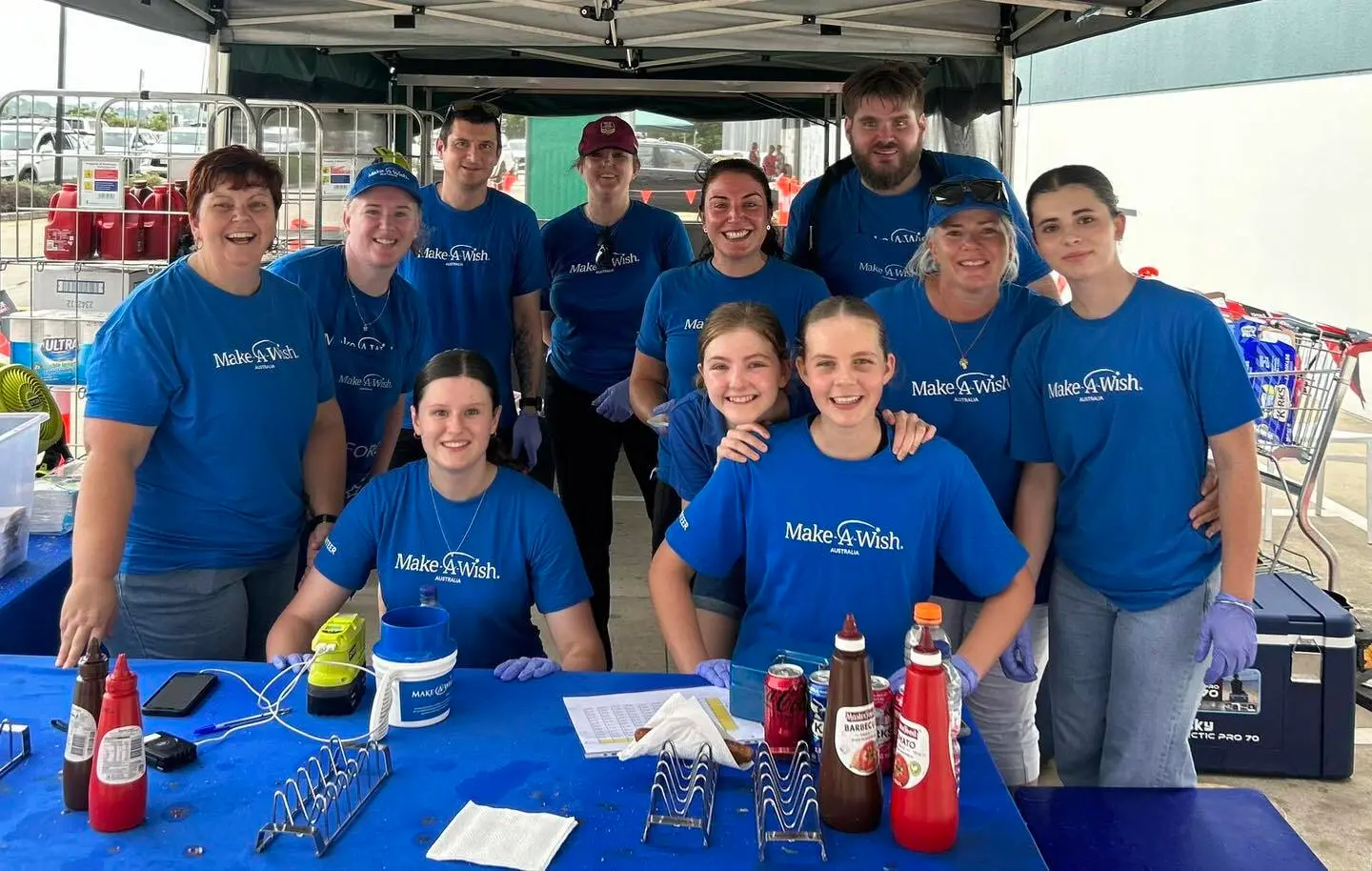 Eleven Make-A-Wish volunteers pose together under a tent in the parking lot of a Bunnings Warehouse. They are all wearing blue Make-A-Wish t-shirts and smiling at the camera. Condiments sit on the table in front of them, ready to be used in the day's sausage sizzle fundraiser.