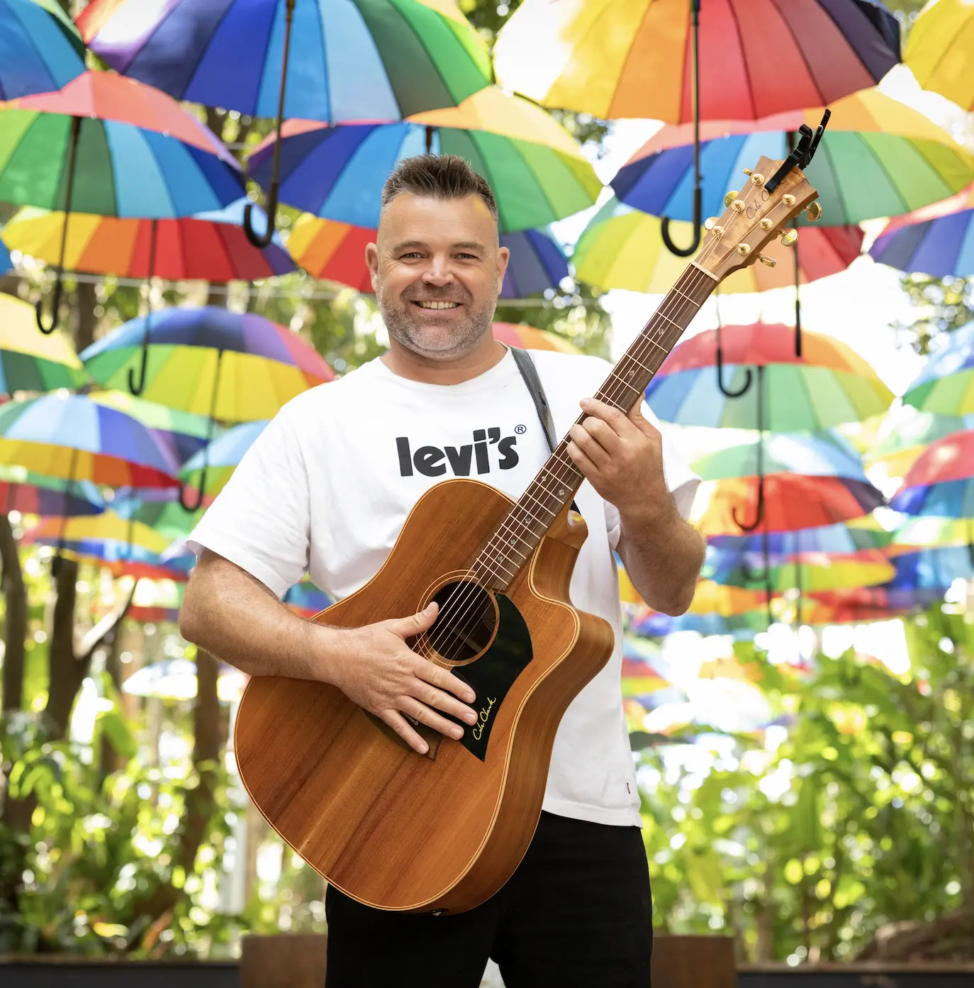 Man with guitar stands in front of rainbow umbrellas
