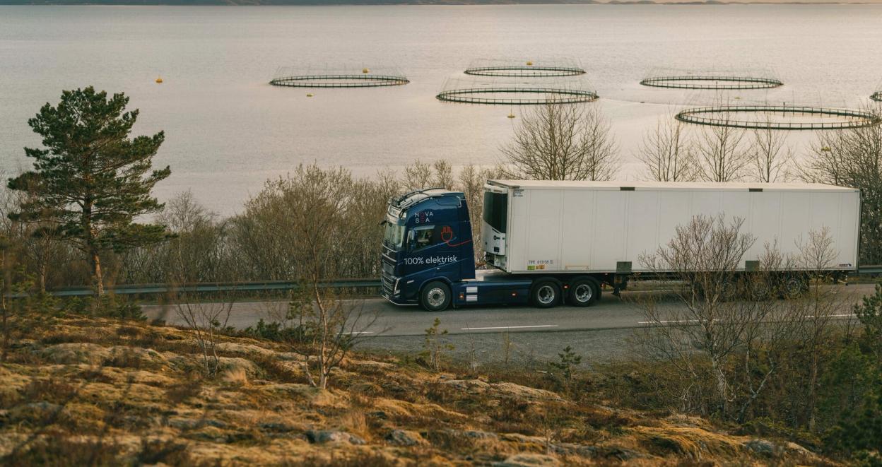 En lastebilde kjører langs landevei, med et oppdrettsanlegg i bakgrunnen