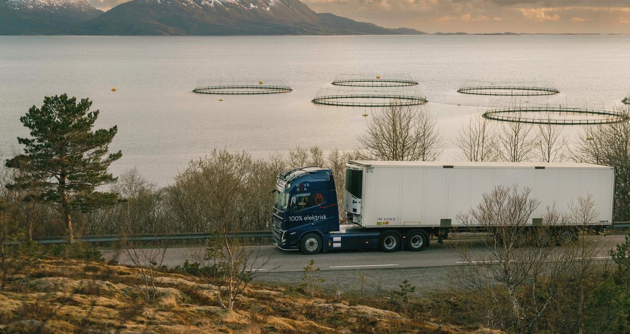 En lastebilde kjører langs landevei, med et oppdrettsanlegg i bakgrunnen