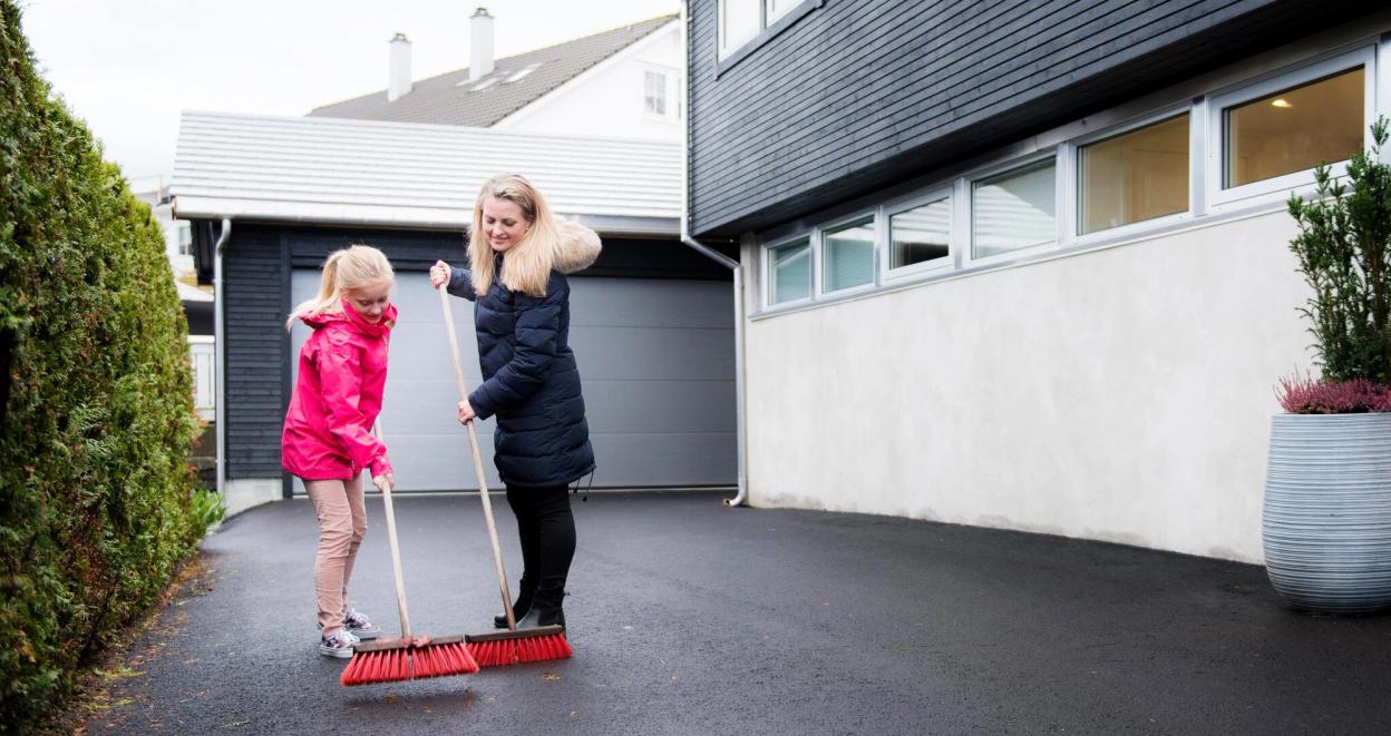 Mor og datter feier grus og løv fra parkeringsplassen ved huset.