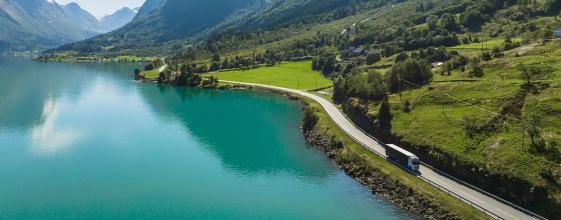 Lastebil kjører langs en distriktsvei i et frodig fjell- og fjordlandskap