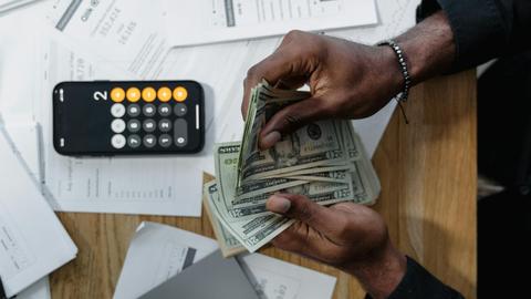 Person counting dollar bills over documents with a smartphone calculator on the desk.