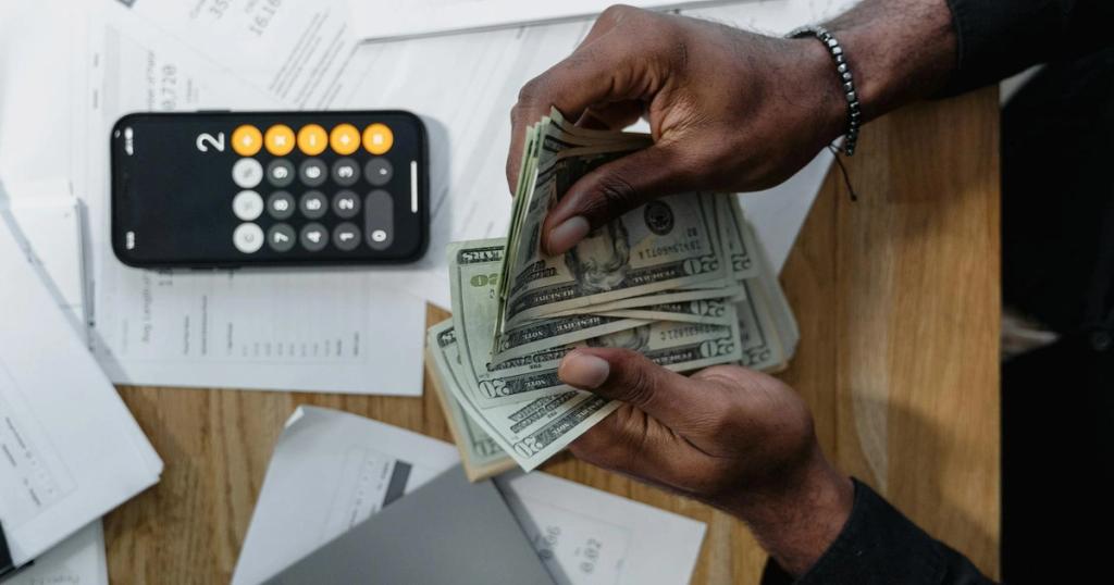 Person counting dollar bills over documents with a smartphone calculator on the desk.