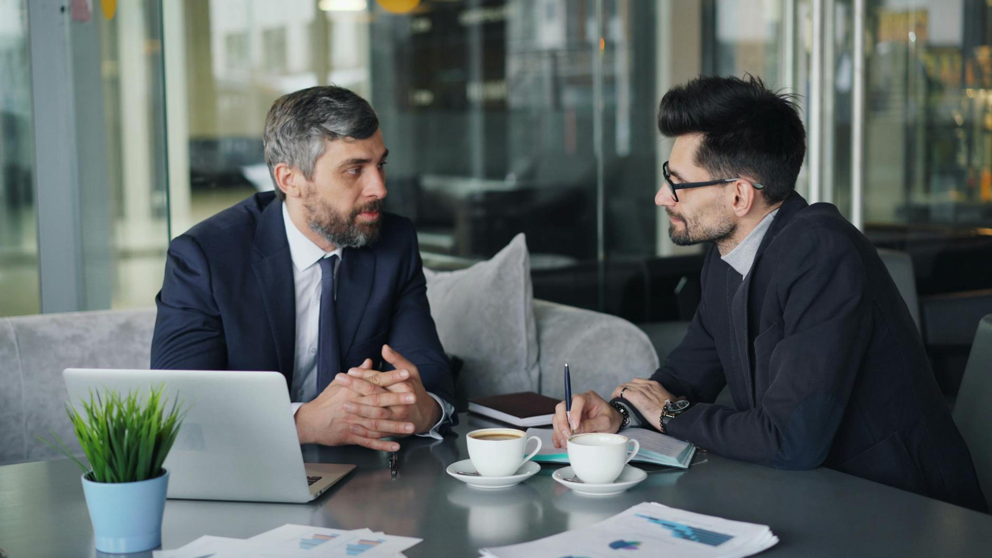 Two businessmen having a meeting with laptops, papers, and coffee at a modern office.