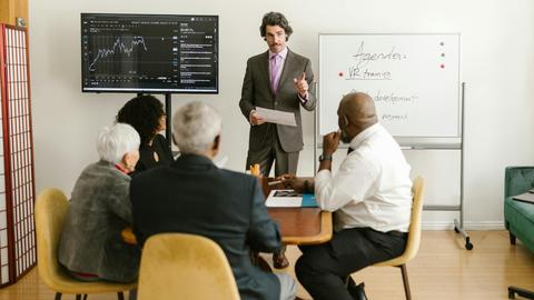 A corporate professional presents market data during a team meeting in an office setting.