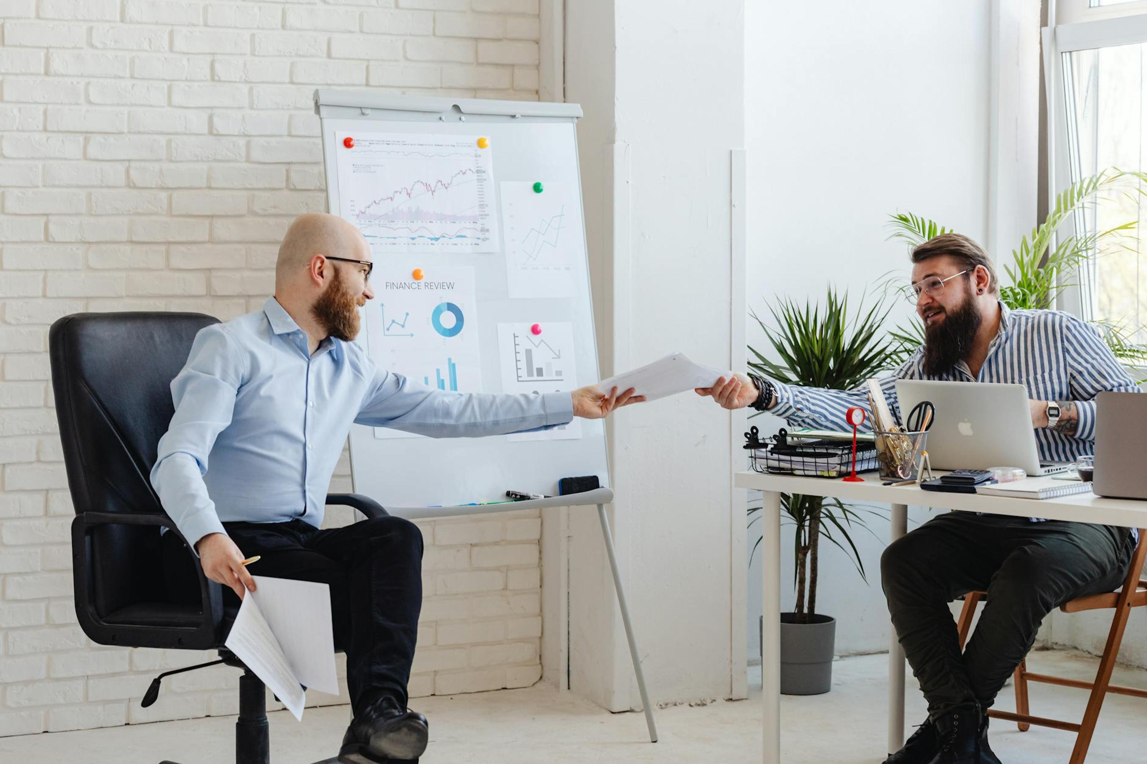Two business professionals exchanging documents in a modern office setting with charts and graphs.