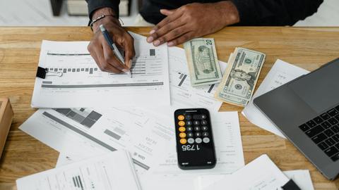 Man working on financial reports with calculator, money, and laptop on a desk.