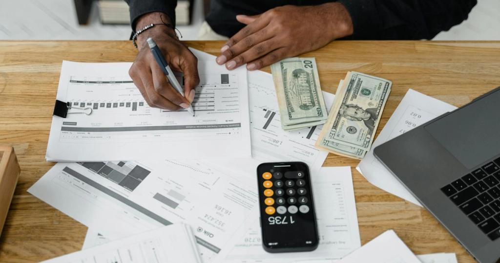 Man working on financial reports with calculator, money, and laptop on a desk.