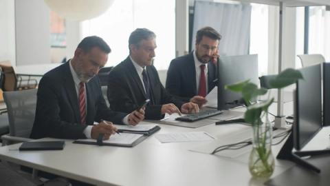Three men in suits collaborating at a modern office desk with computers and documents.