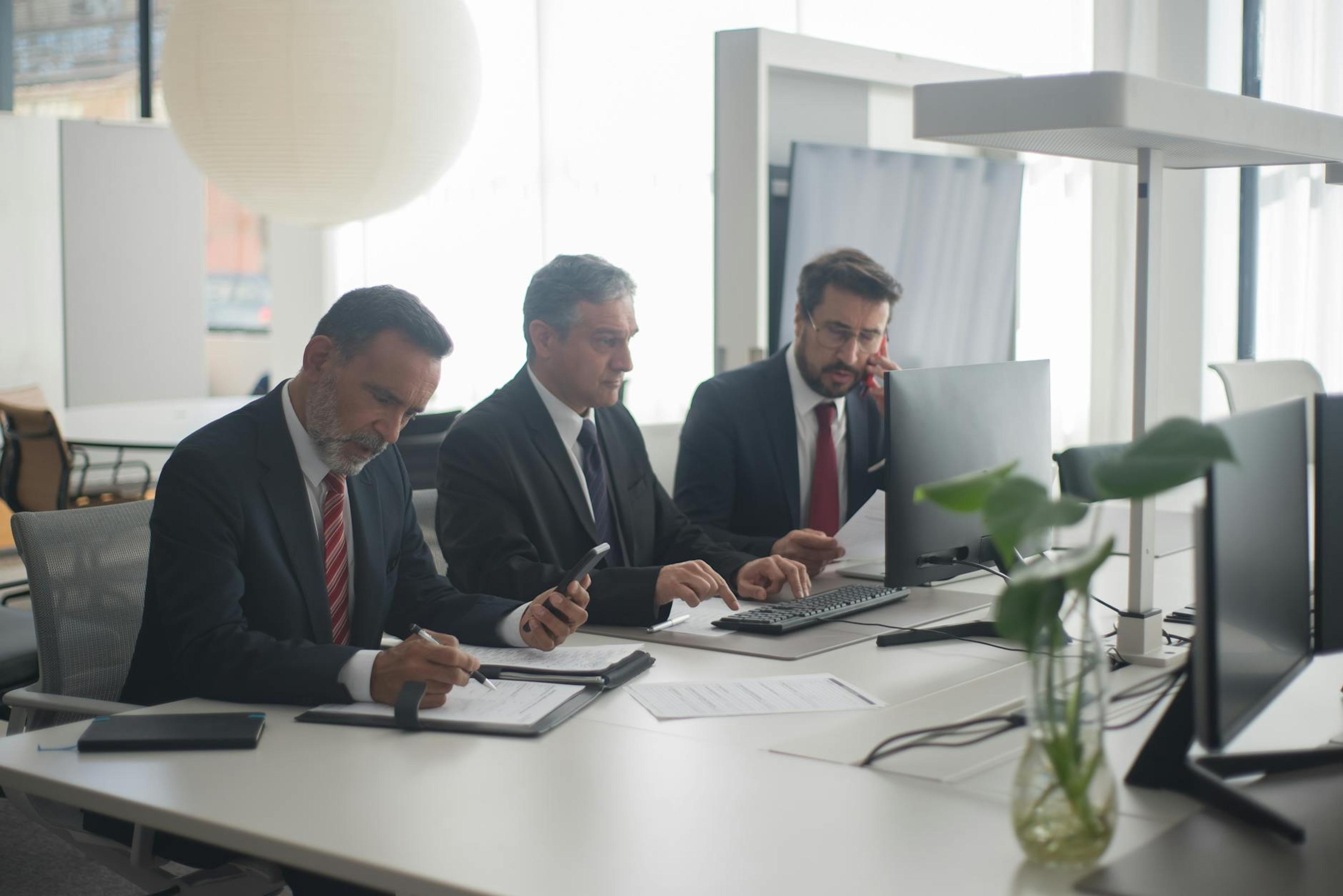 Three men in suits collaborating at a modern office desk with computers and documents.