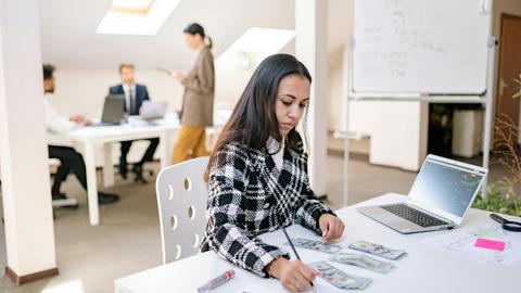 Focused businesswoman reviewing financial documents and currency in a modern office setting.