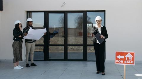 Real estate professionals with hard hats inspect a property for sale. Outdoor meeting with blueprints.