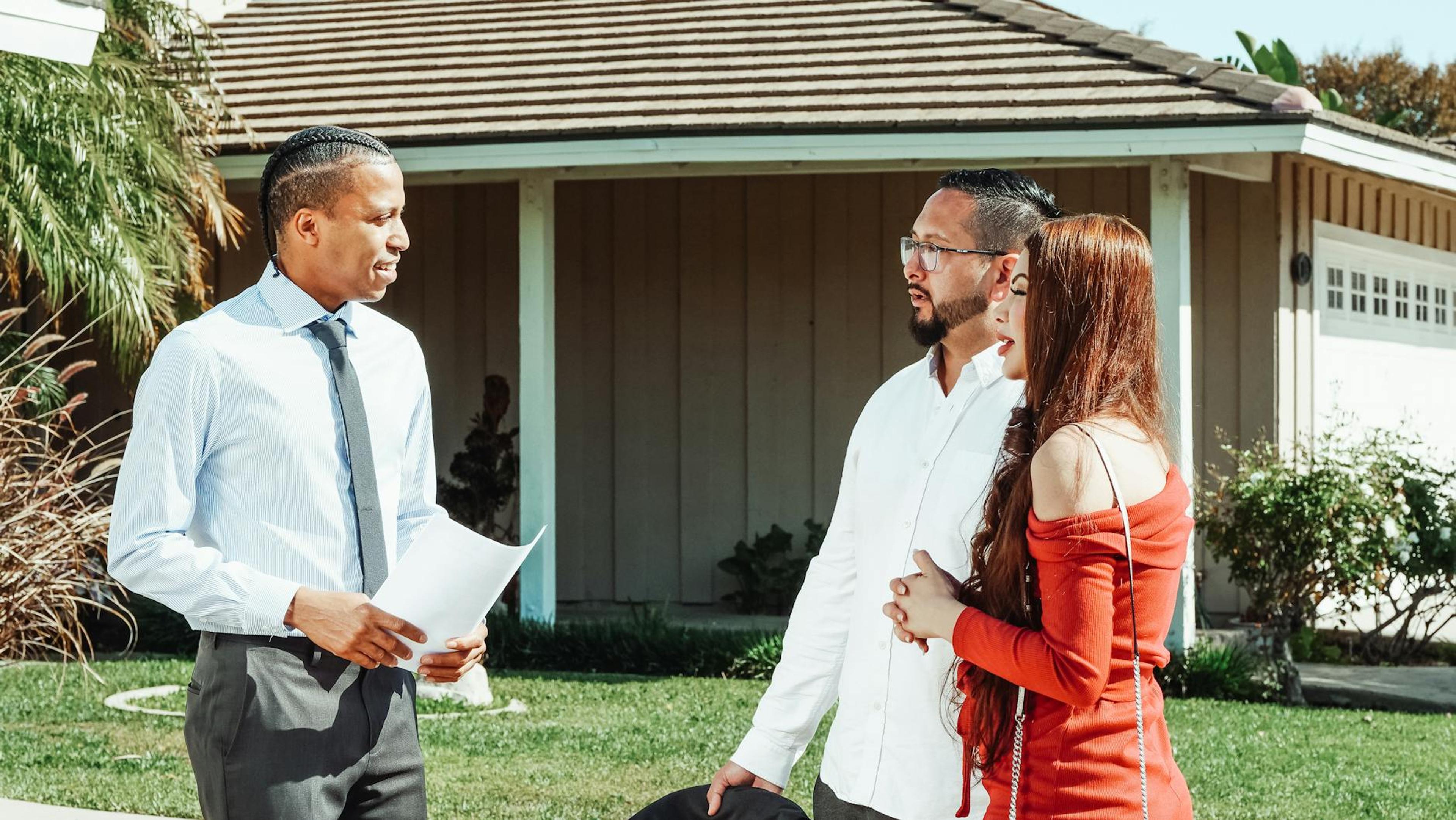 A real estate agent discusses property details with a couple outside a suburban home.