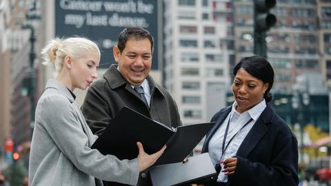 Young well dressed woman sharing folder with multiracial colleagues while talking in town in daytime