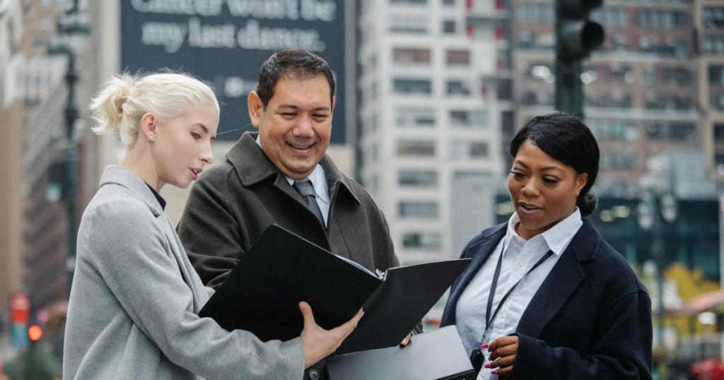 Young well dressed woman sharing folder with multiracial colleagues while talking in town in daytime